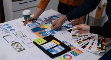 Team of coworkers reviewing printed design mockups and charts on a table in an office, collaborating on branding or marketing strategy with coffee cups and stationery.